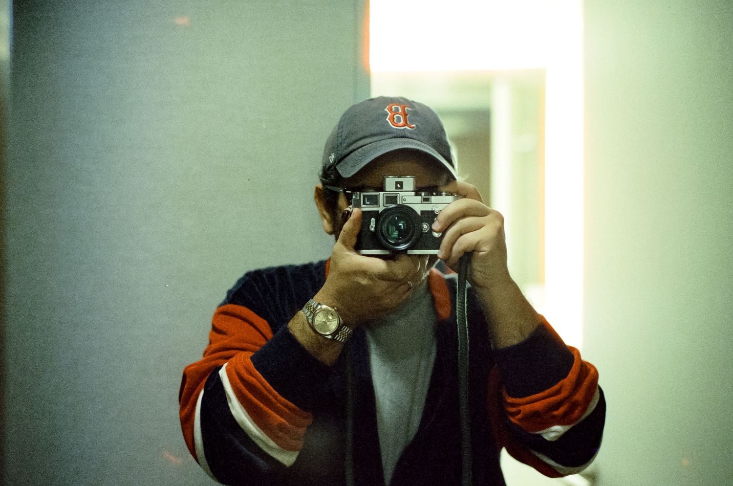 Someone taking a mirror selfie with a vintage camera while wearing a baseball cap and jersey.