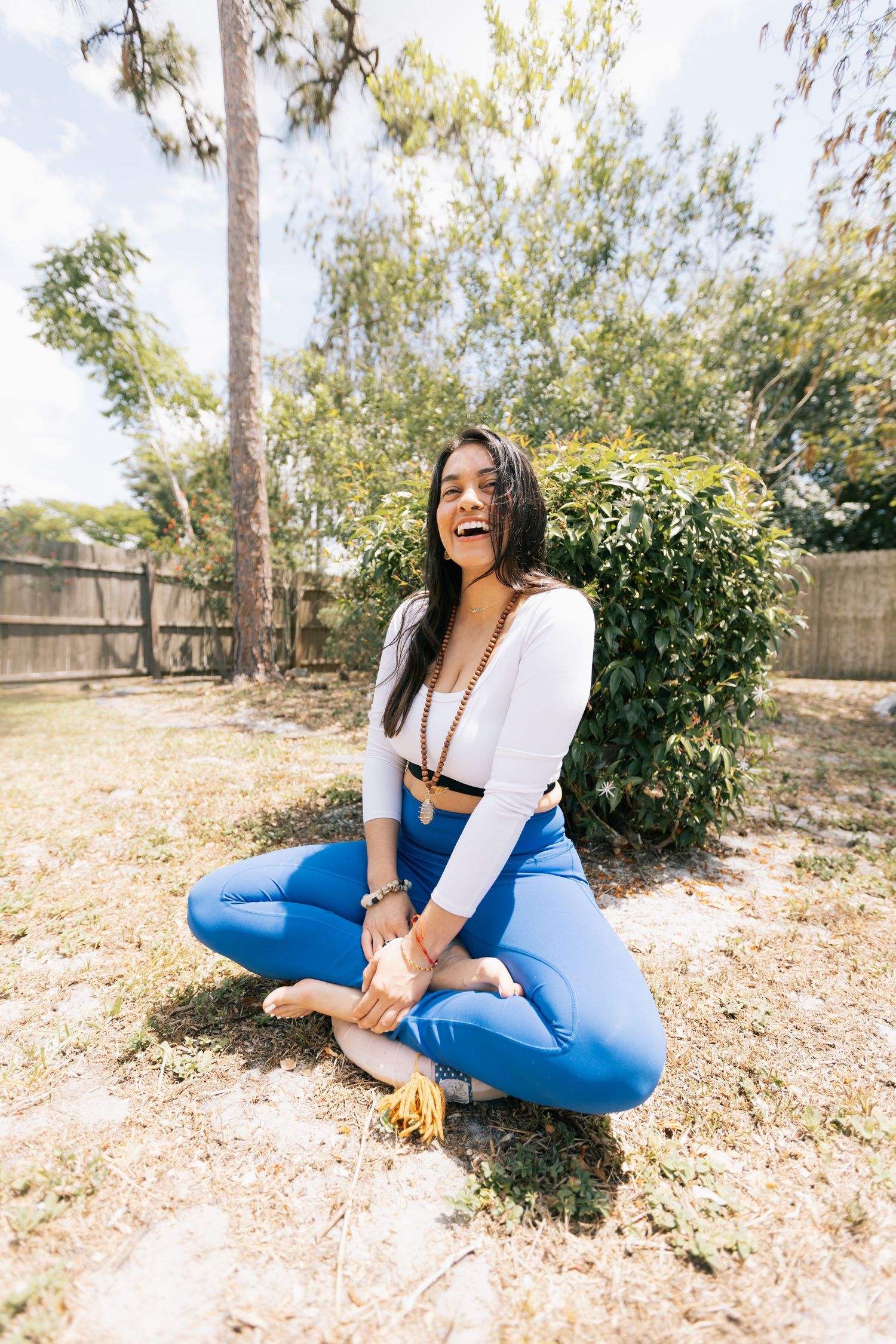 A joyful woman in blue leggings and a white top sits cross-legged and laughing on a sunny backyard lawn near a wooden fence.