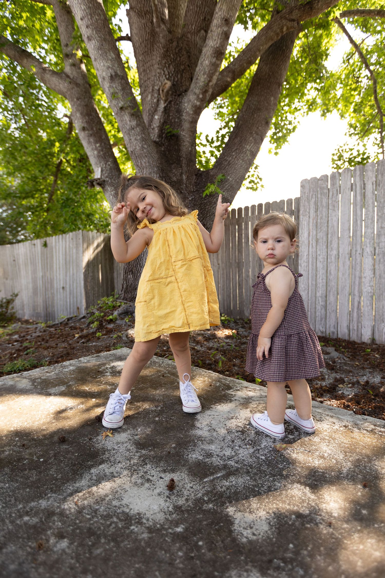 Two young girls in a yellow dress and a mauve dress pose playfully on a concrete patio beneath a large shady tree.