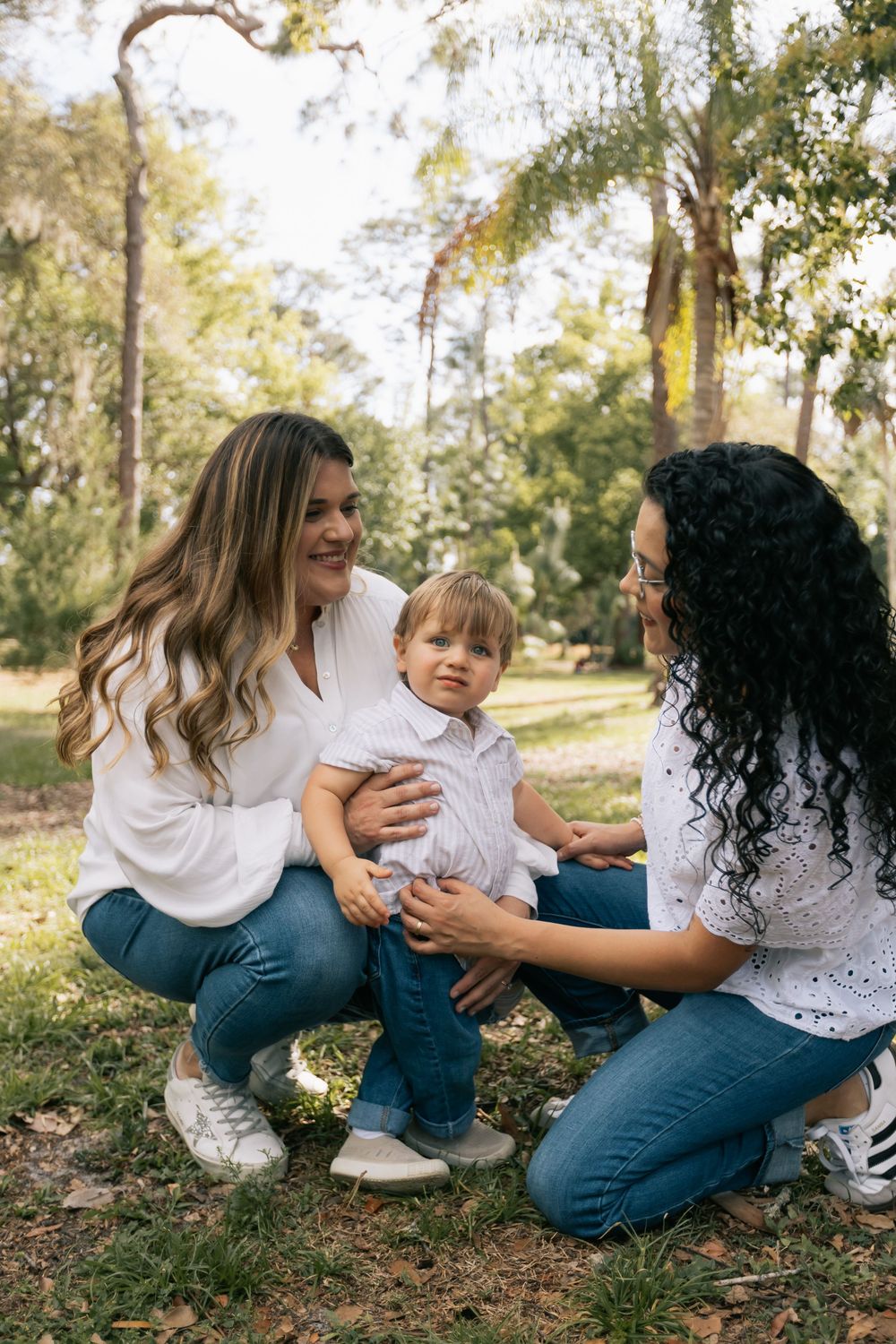 Two smiling women in jeans and white tops kneel on the grass hugging a toddler boy in a sunny park.