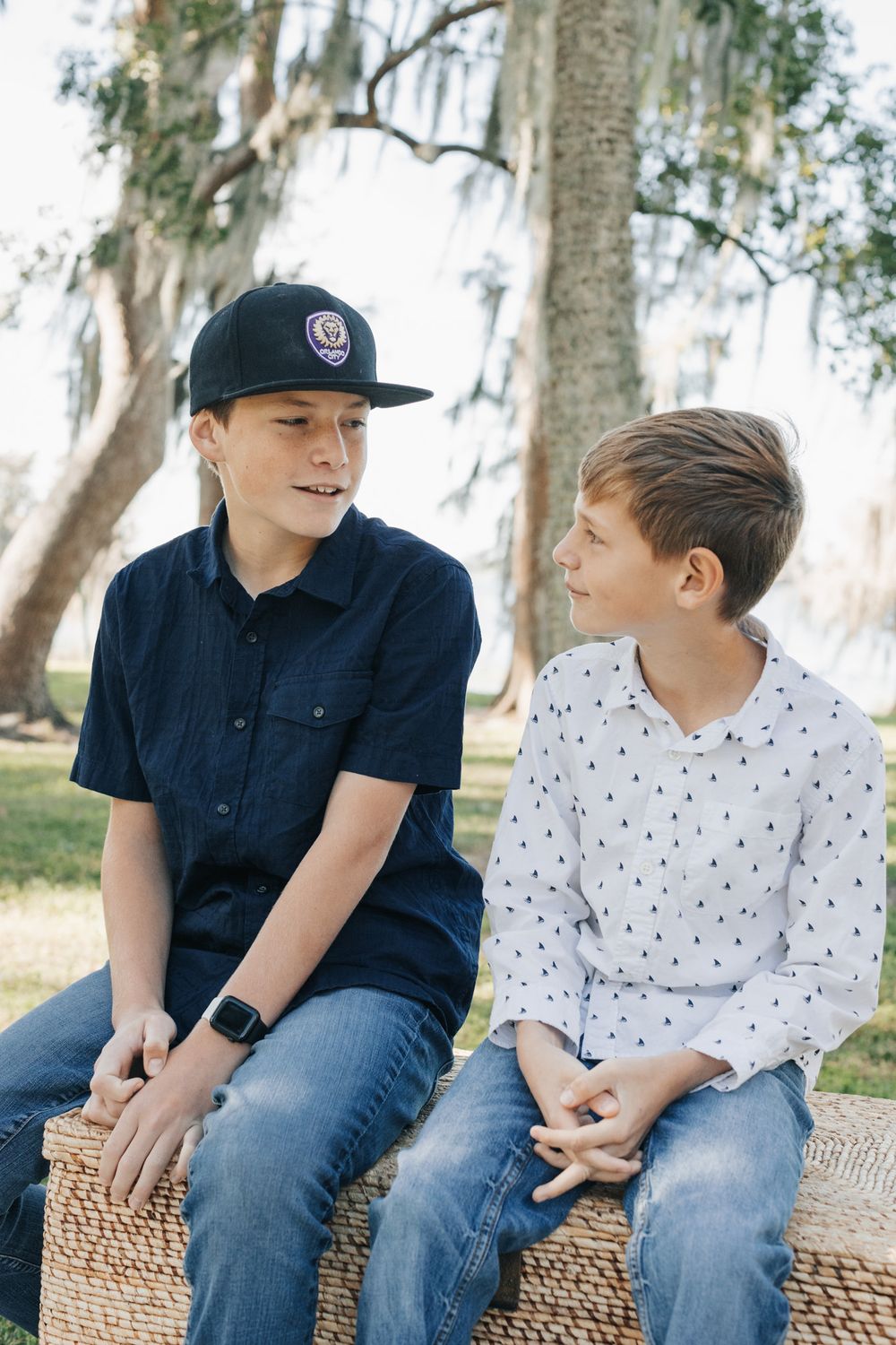Two boys sit on a woven bench outdoors, one in a black cap and navy shirt, chatting beneath mossy oak trees.