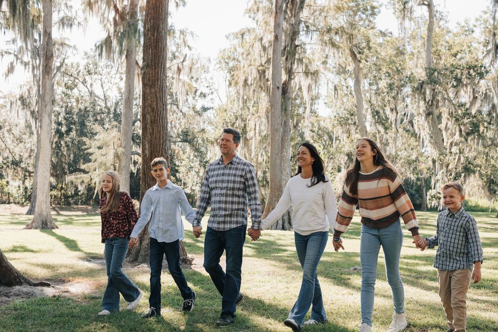 A family of six holds hands and walks together beneath tall oak trees draped in Spanish moss on a bright day.