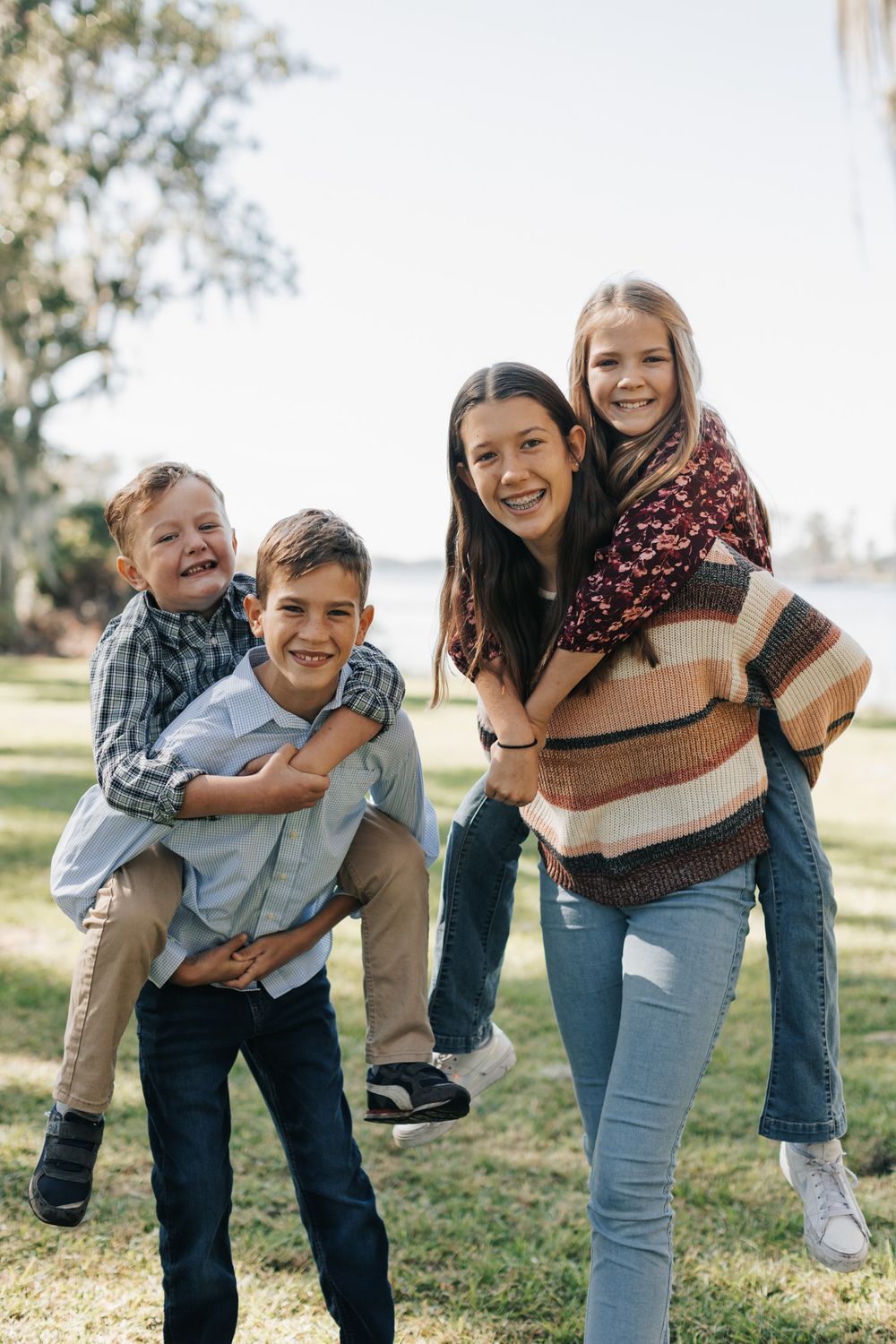 A teenage girl and a boy give two smiling younger children piggyback rides on a sunny lawn by the water.