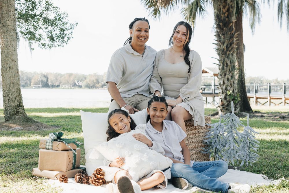 A family of four poses on a picnic blanket with pinecones and a gift box beside a lake and palm trees.