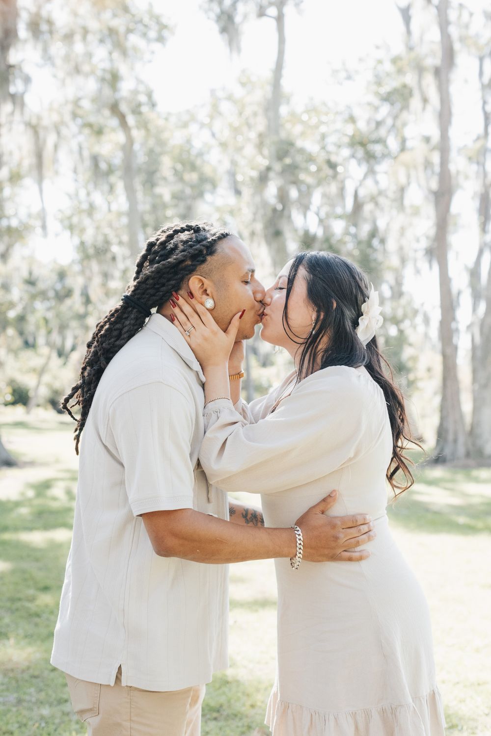 A couple in neutral outfits share a tender kiss in a sunlit park lined with moss-draped trees.