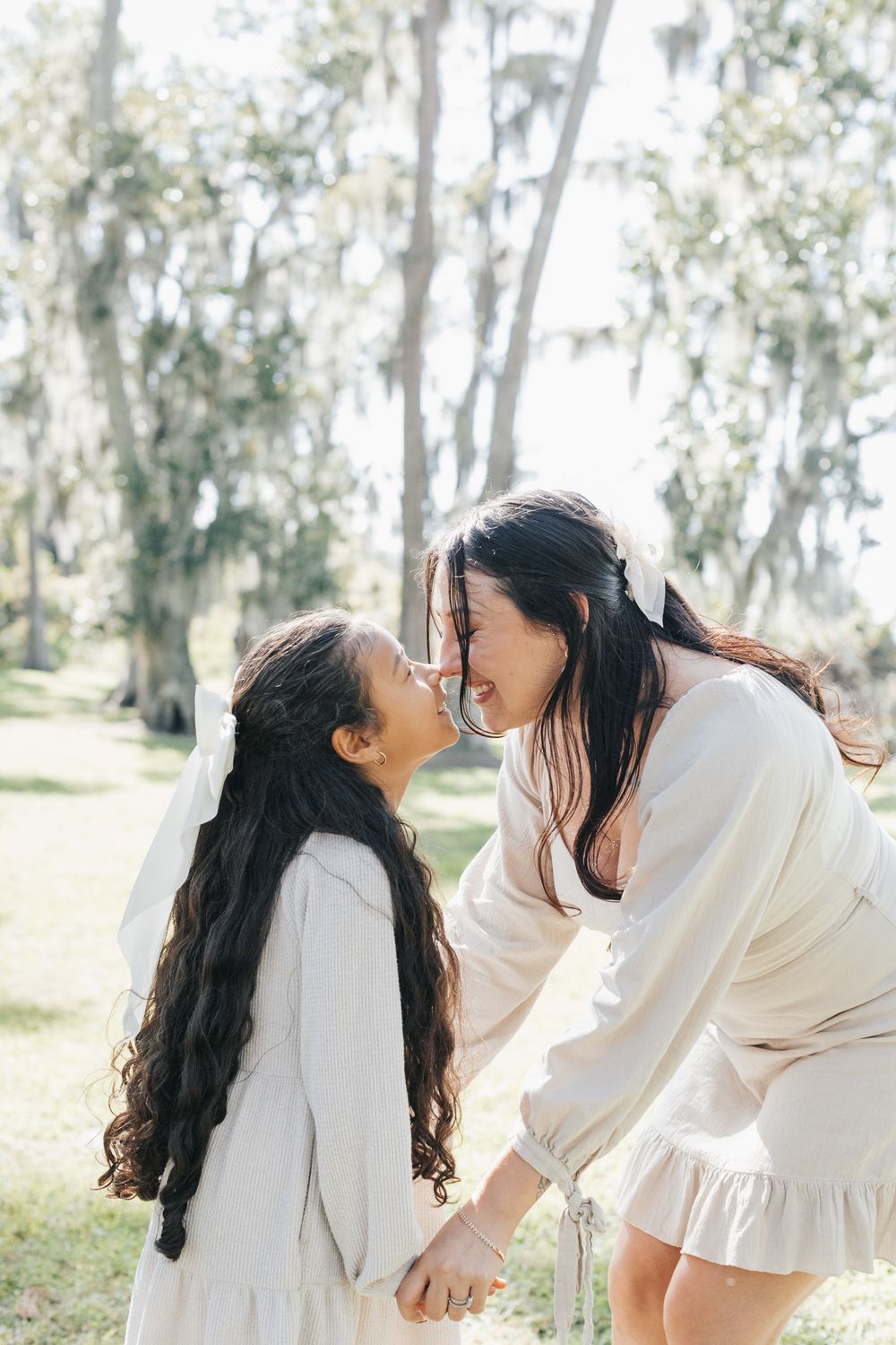 A mother and daughter with long dark hair touch noses and hold hands in a sunlit park with tall trees.