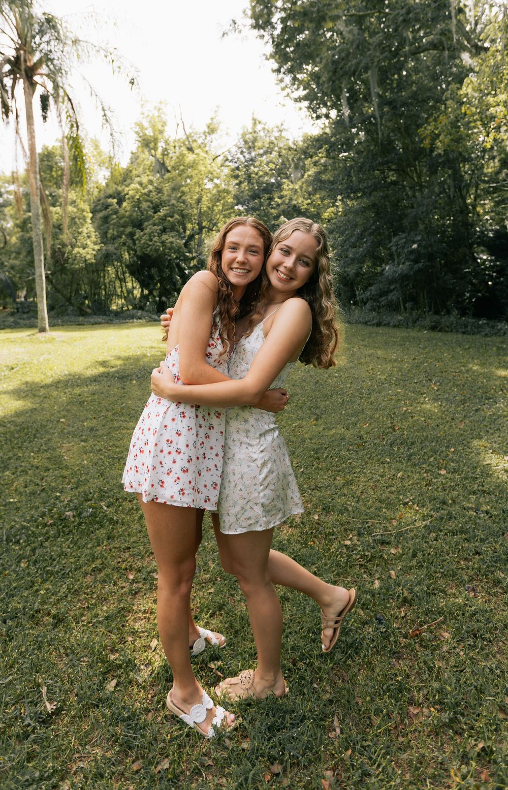 Two smiling girls in floral summer dresses hug each other on a sunny grassy lawn surrounded by tall green trees.
