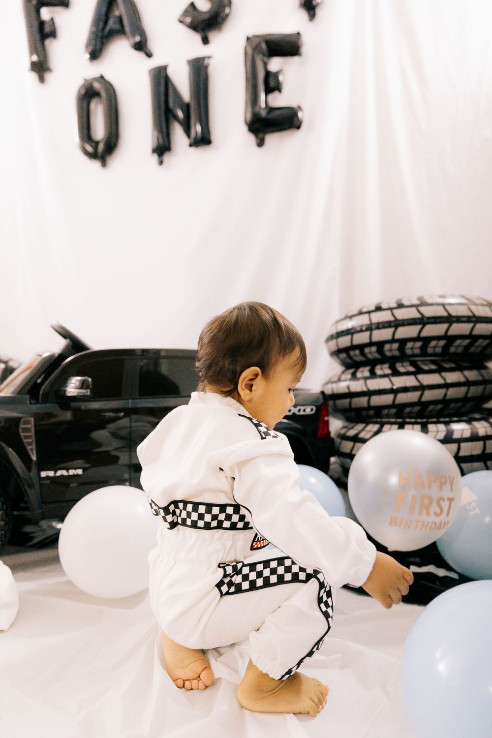 A barefoot toddler in a white checkered racing suit sits by balloons and tires at a race car themed first birthday party.