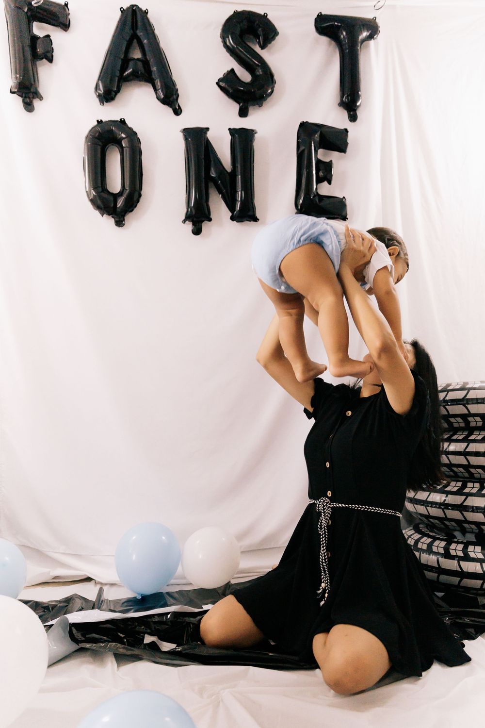A woman in a black dress lifts a baby into the air beneath black Fast One balloon letters at a racing themed birthday.