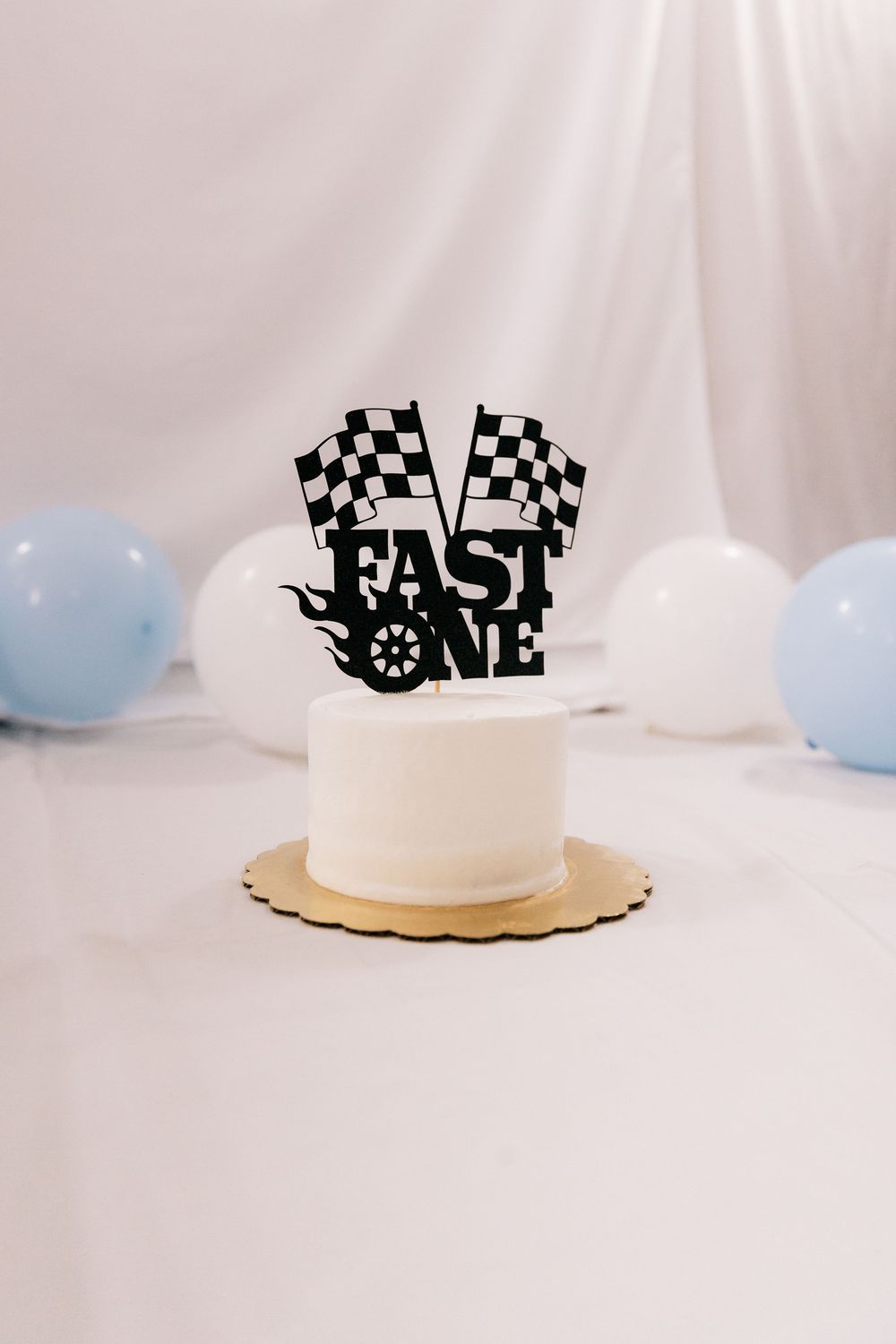 A white Fast One birthday cake with checkered flag topper sits on a table surrounded by blue and white balloons.