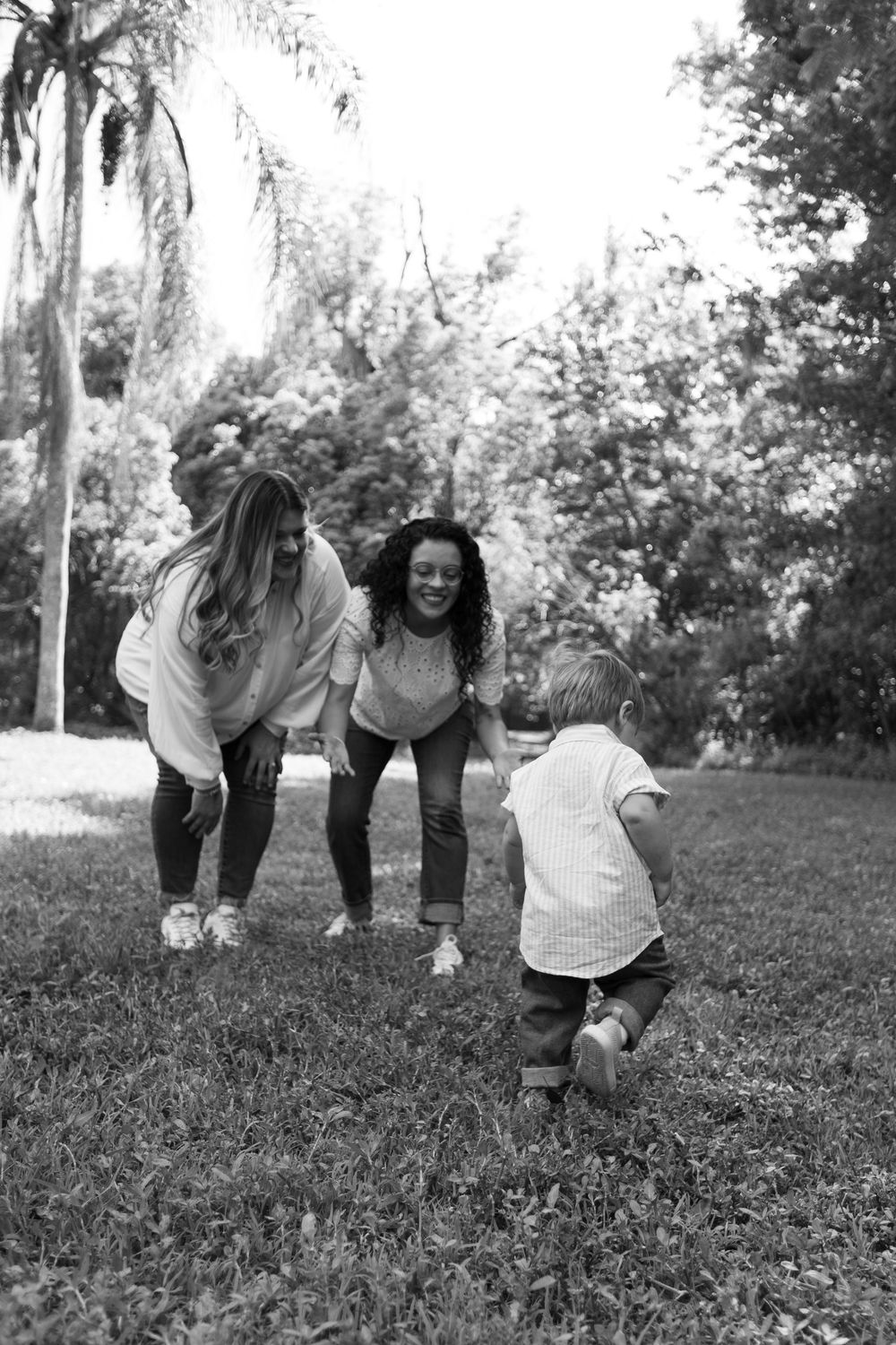 Black and white photo of two women crouching with open arms as a toddler walks toward them across a grassy park.