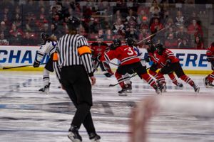 Hockey players in red jerseys face off on the ice as a referee in black and white stripes observes the game.