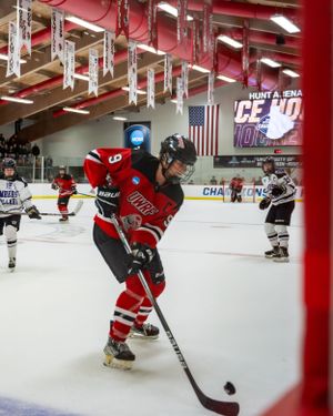 Hockey player in red jersey controls puck during game with American flag and championship banners visible in background.