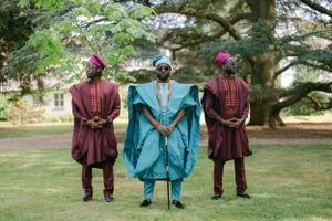 Three people stand in traditional African formal wear with flowing robes in burgundy and bright blue.