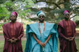 Three figures in matching African formal attire pose in garden, featuring burgundy and turquoise agbada robes.