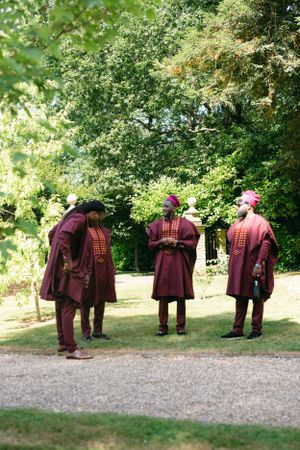 Three figures in burgundy traditional outfits and hats standing on a garden path surrounded by lush greenery.