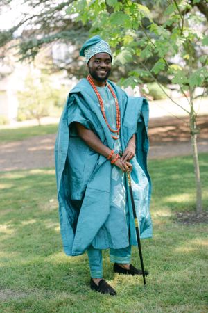 Person in bright turquoise traditional African clothing holding walking stick poses in garden setting.
