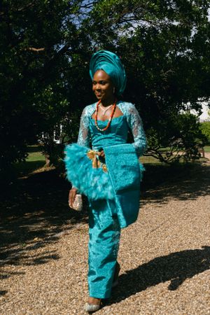 Woman in vibrant turquoise traditional dress and headwrap poses on gravel path.