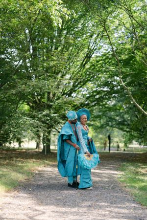 A couple in traditional teal African clothing walk together on a tree-lined path.