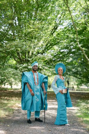A couple in matching turquoise Nigerian formal wear pose elegantly on a forest path.