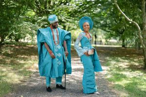A couple in coordinated teal African attire stand together in a sunlit park setting.