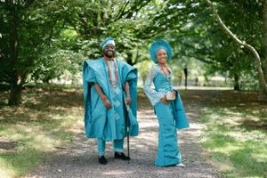 A couple in matching teal Nigerian outfits pose together on a wooded path.