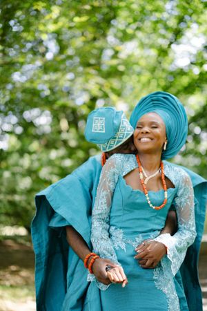 Woman in elegant turquoise traditional Nigerian gele and lace dress adorned with coral beads in a garden setting.