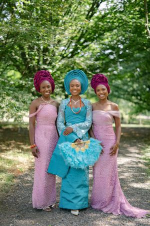 Three elegantly dressed women in pink and turquoise gowns with matching headwraps pose on garden path.