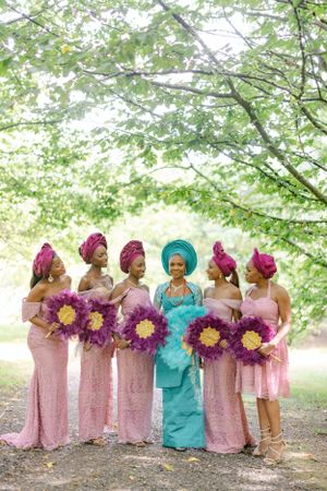 Wedding party group in pink dresses and head wraps poses with purple bouquets under trees.