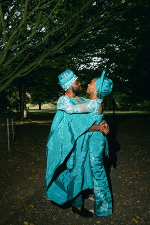 Two people in vibrant turquoise traditional Nigerian attire embrace under trees in a park.