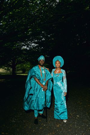 A couple in matching turquoise Nigerian formal wear poses in a park setting.