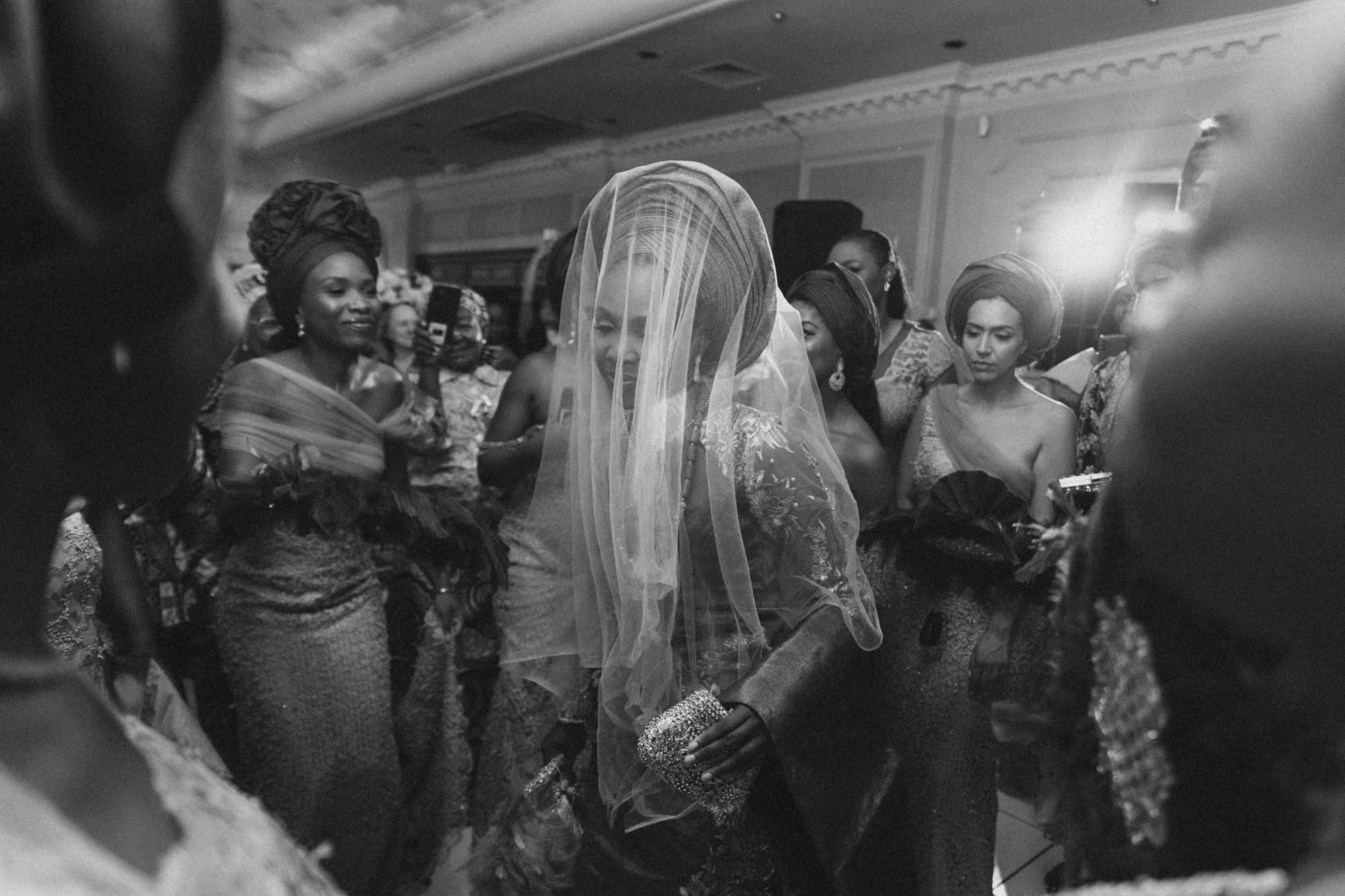 A black and white photo captures a festive celebration with a veiled figure at the center.
