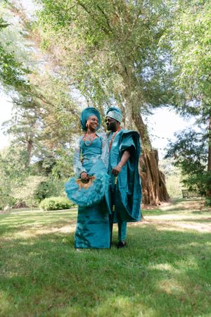 Two people in matching turquoise ceremonial outfits pose together in a garden setting.