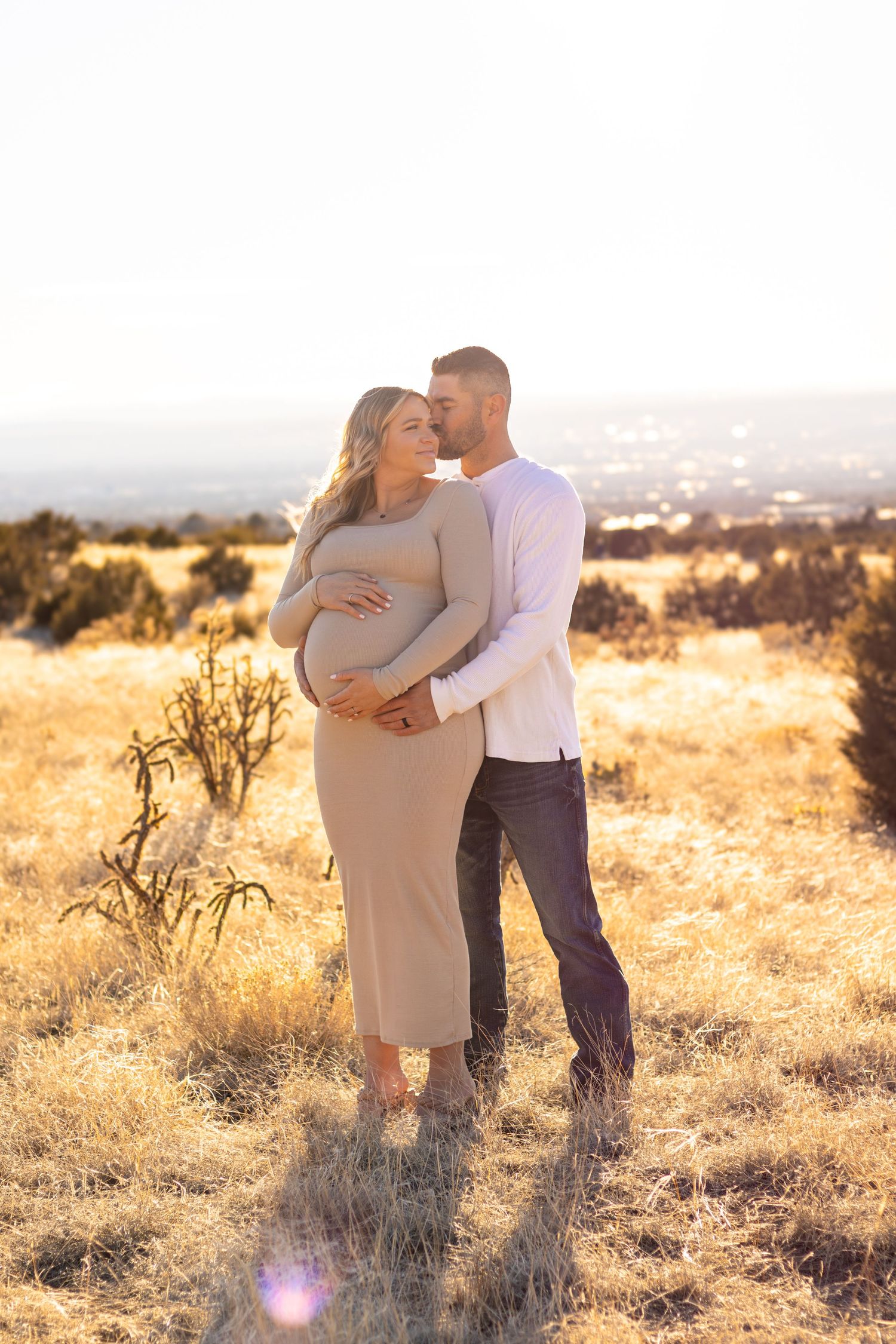 Expectant couple embraces in golden field during sunset maternity photoshoot.