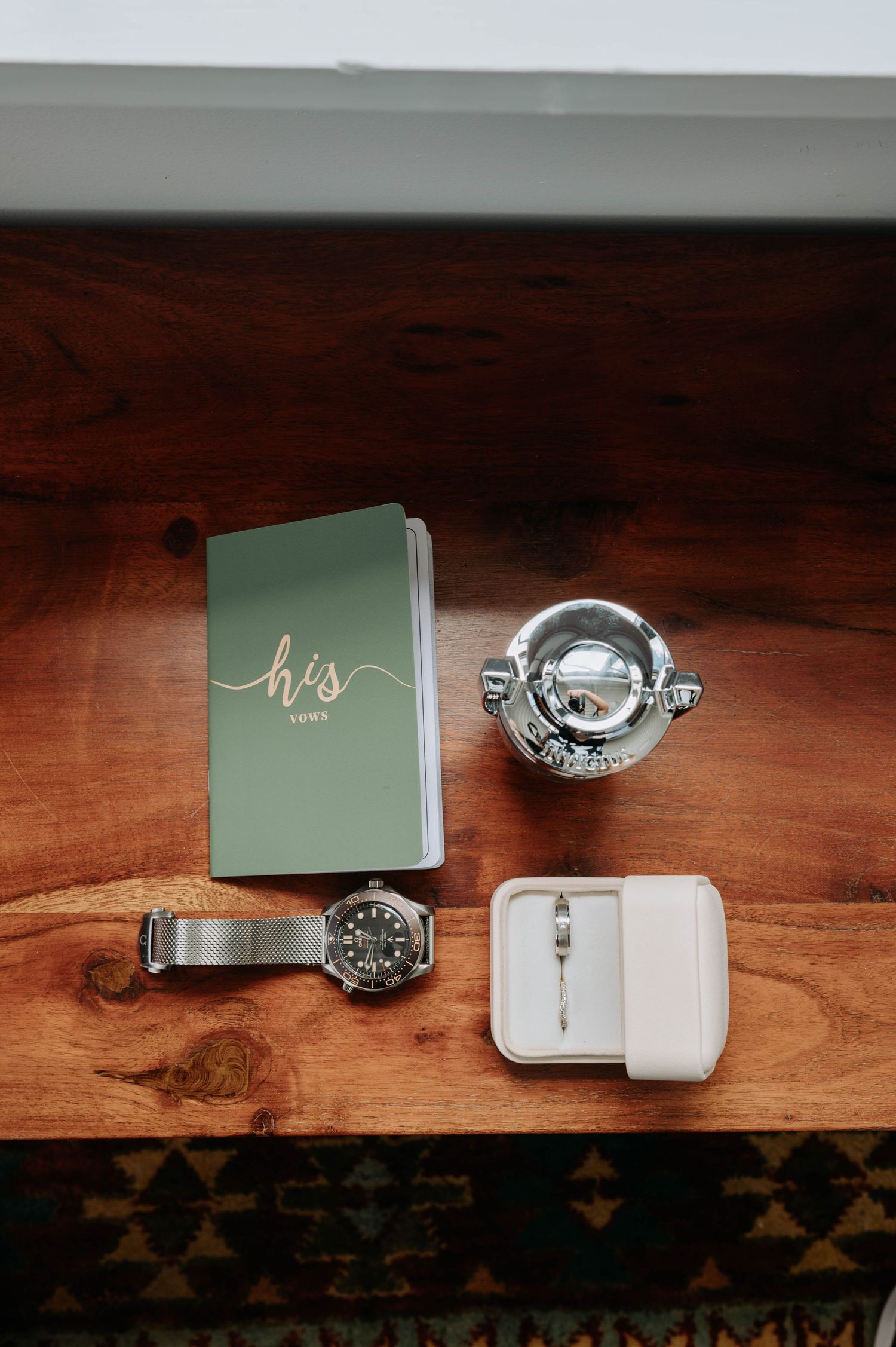 Wedding accessories including vows book, watch, ring box and decorative items on wooden surface.