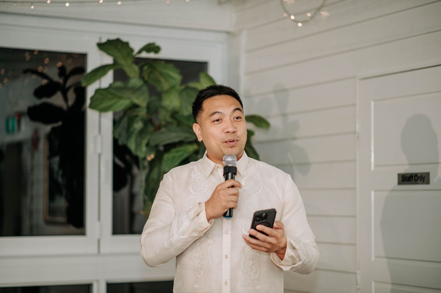 Person in white formal attire speaking into microphone while holding phone during indoor event.