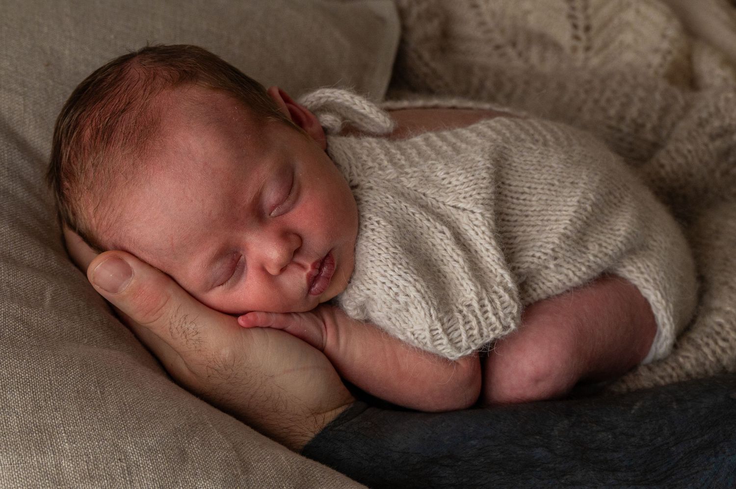 Sleeping newborn baby in white sweater rests on cushioned surface.