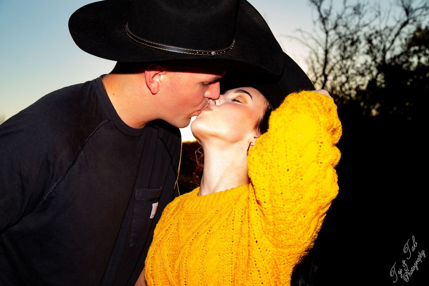 Couple kissing at sunset with one wearing a black cowboy hat and other in bright yellow sweater.