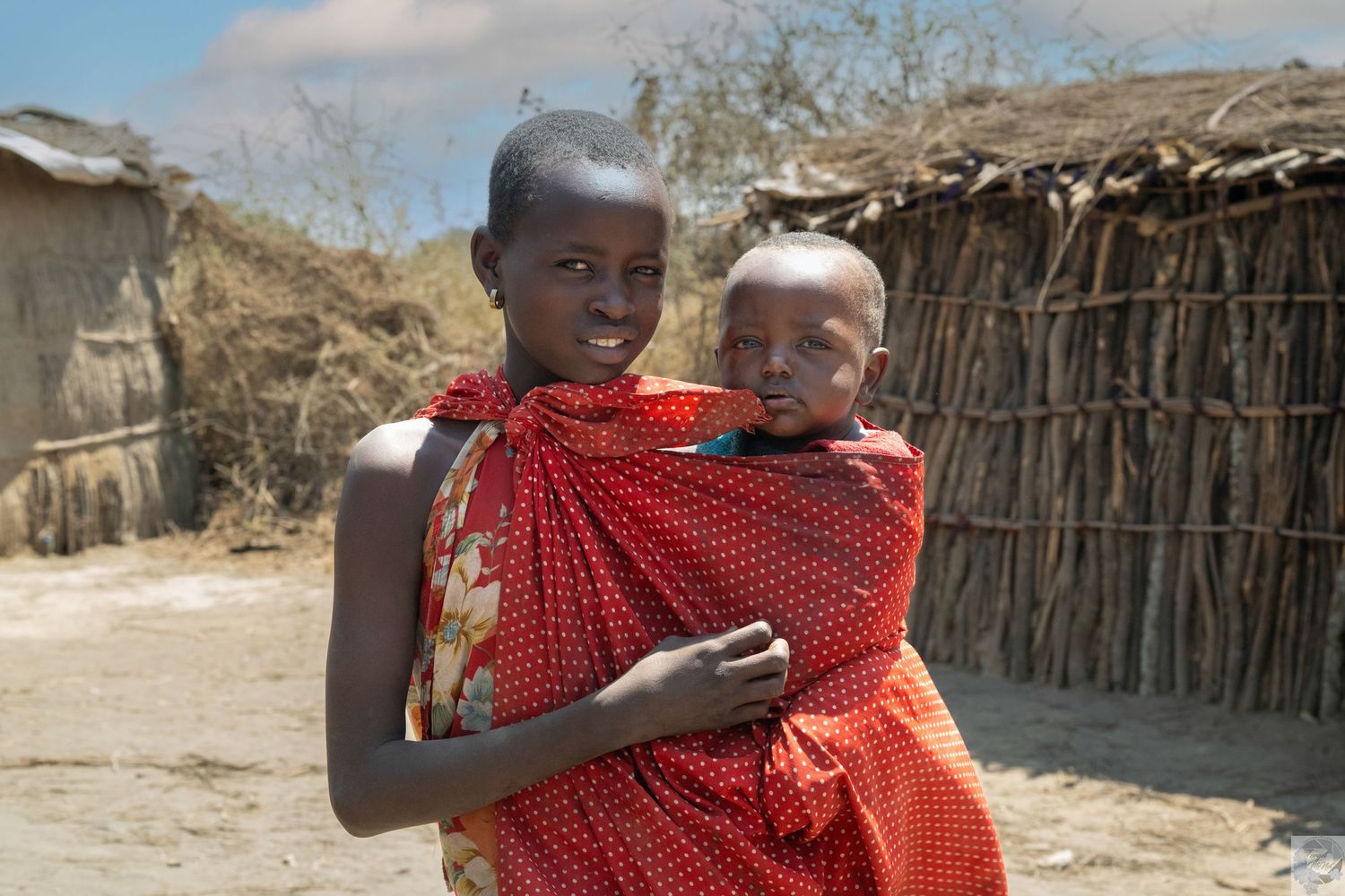 Children in traditional red clothing stand near thatched huts in an African village.