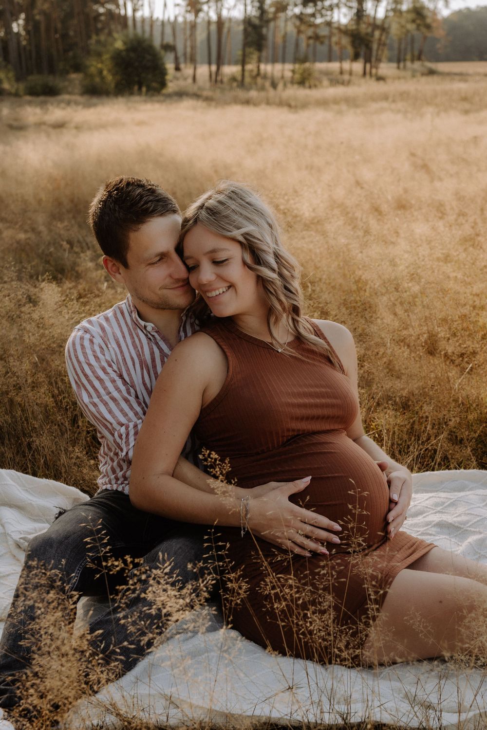 Couple shares intimate moment during maternity photo session on blanket in golden field.