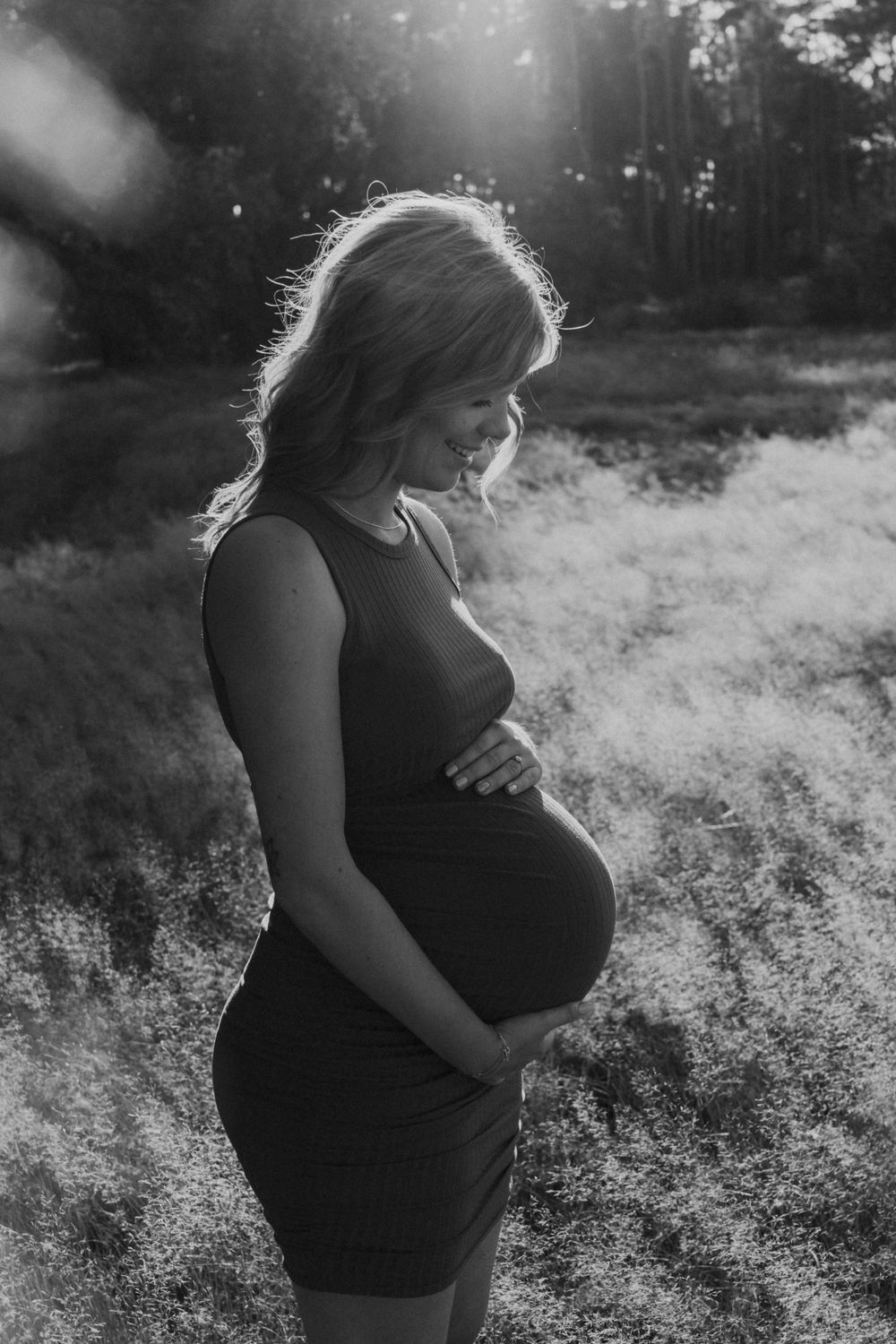 Silhouetted maternity portrait in black dress against sunlit forest backdrop.