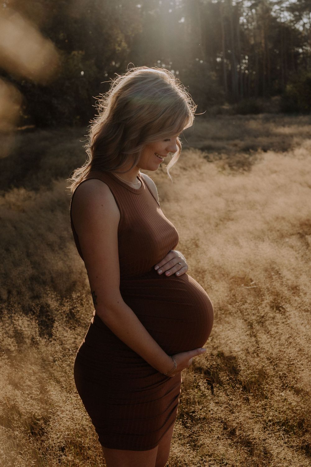 Woman in brown dress poses for maternity photo with sunlit forest backdrop.