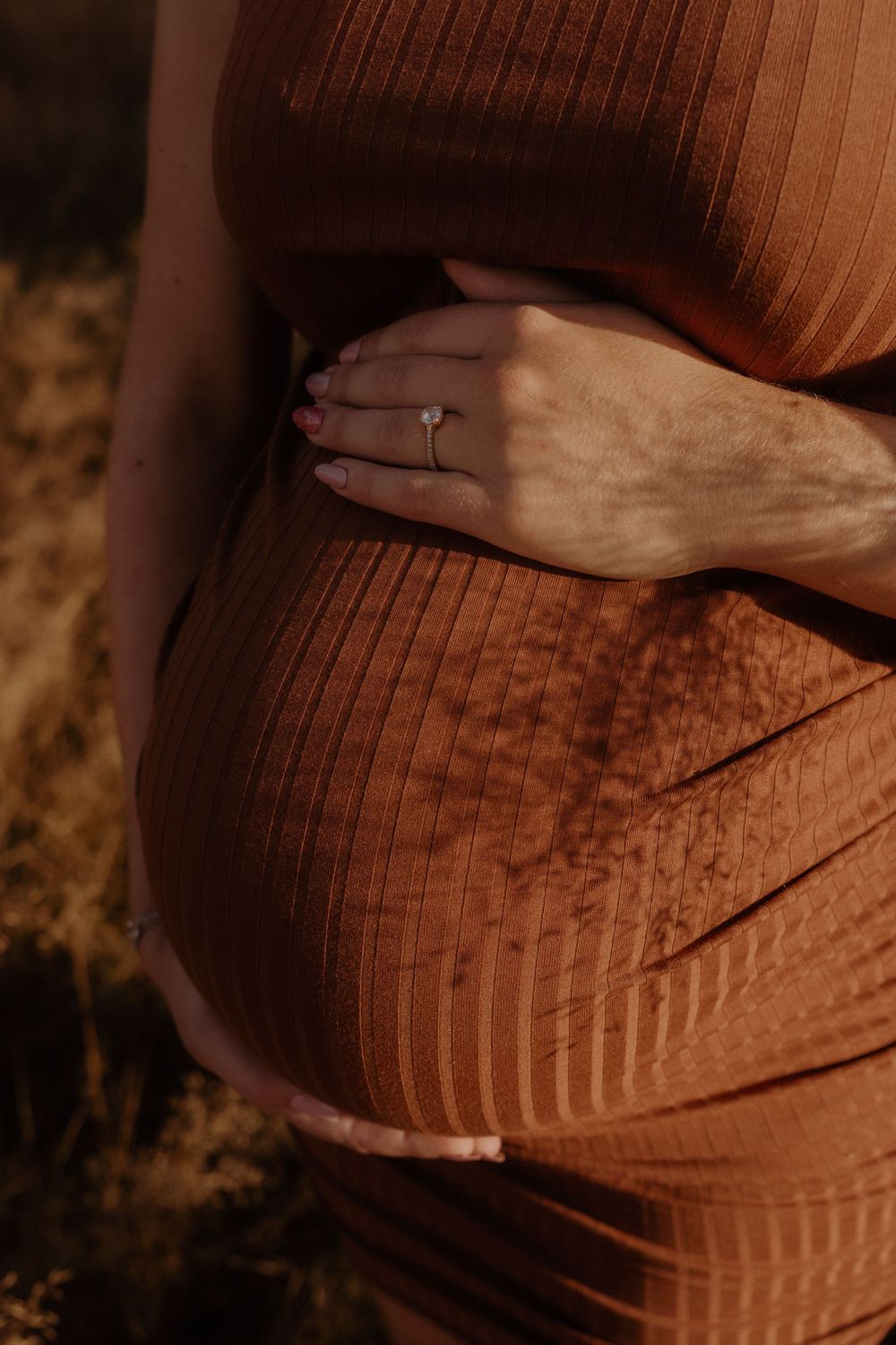 Close-up of hand with engagement ring resting on maternity dress in warm sunlight.