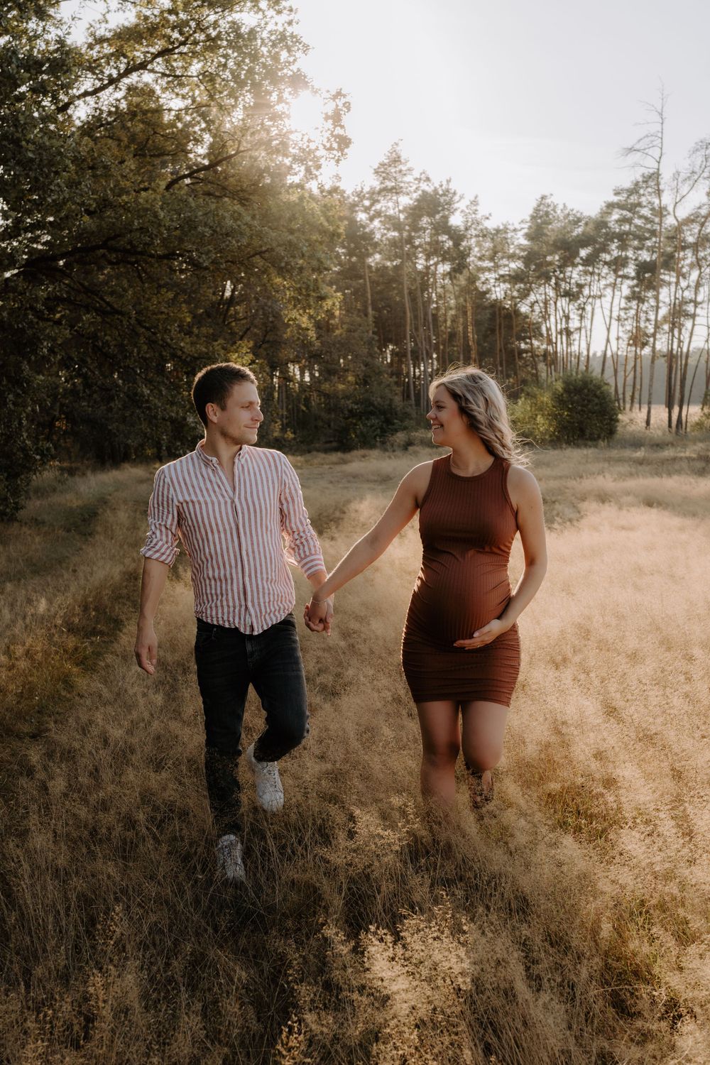 A couple walks hand in hand through a sun-drenched field with tall pine trees in the background.
