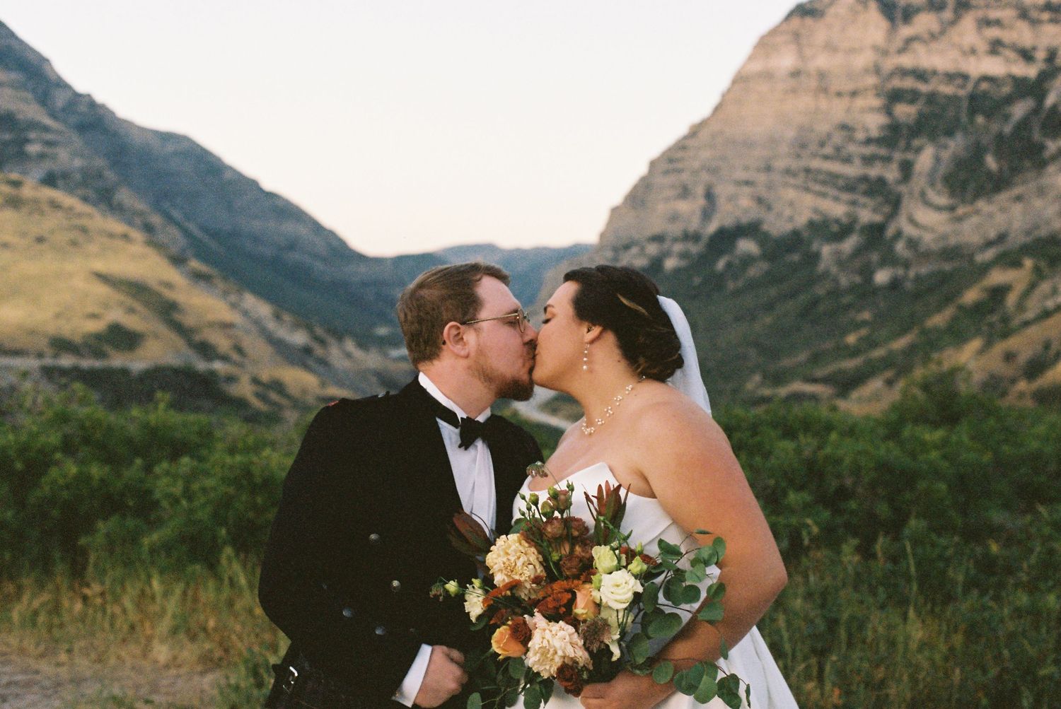 Bride and groom kissing with mountain backdrop, bride holding elegant floral bouquet at outdoor wedding.