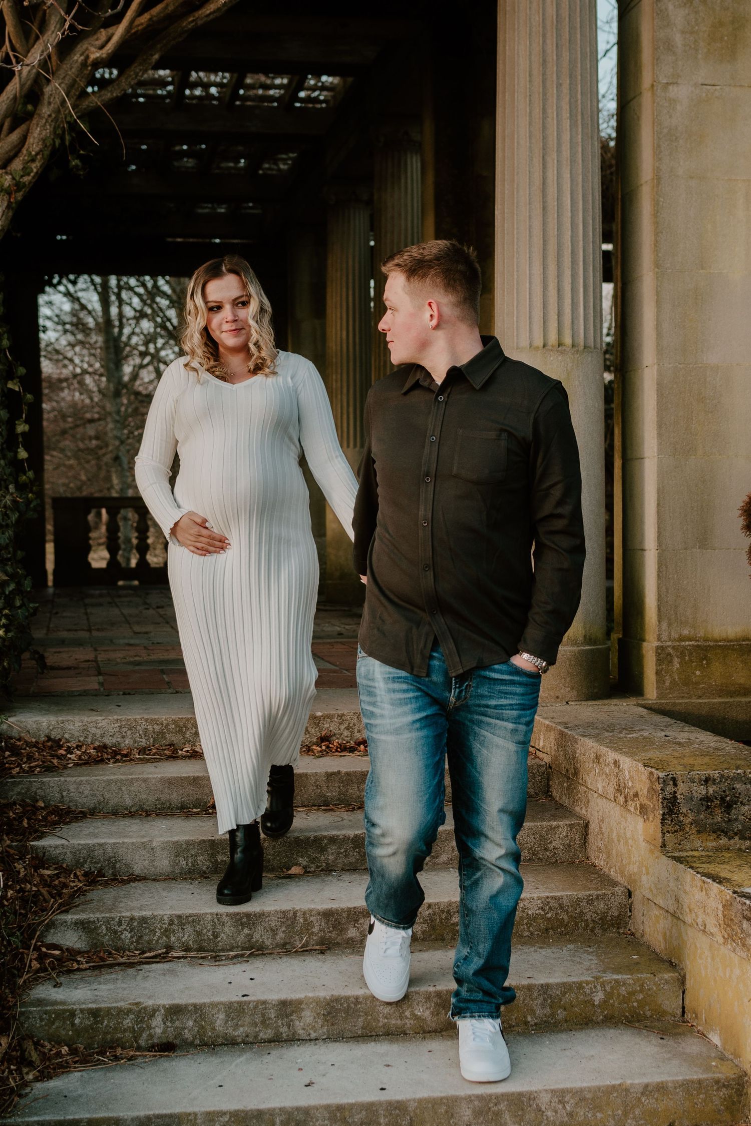 Expecting couple walks down outdoor stone steps together near marble columns at sunset.