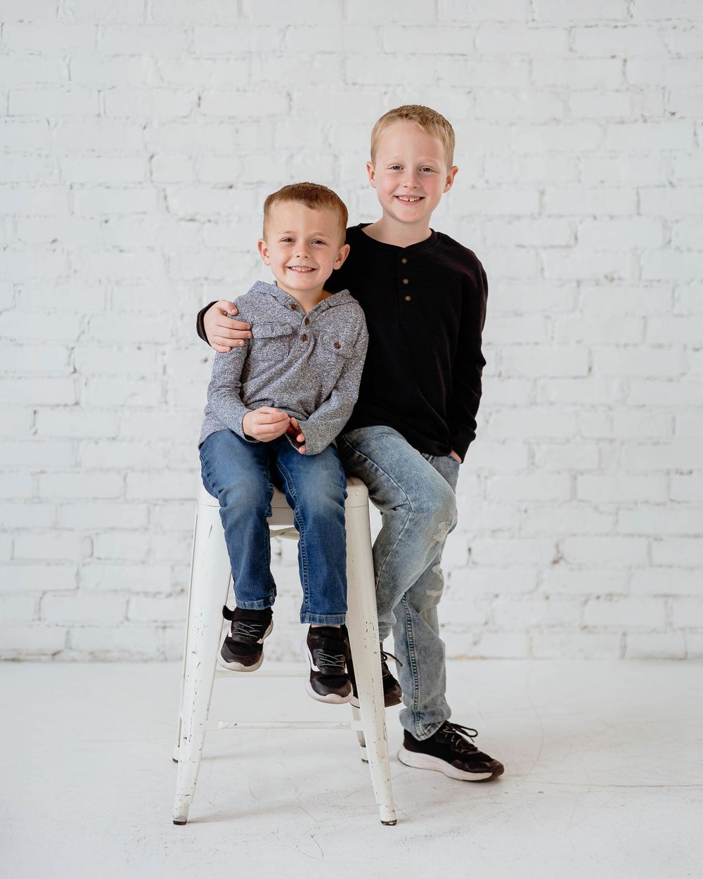 Two children sit together on a white stool against a white brick wall backdrop in a studio setting.