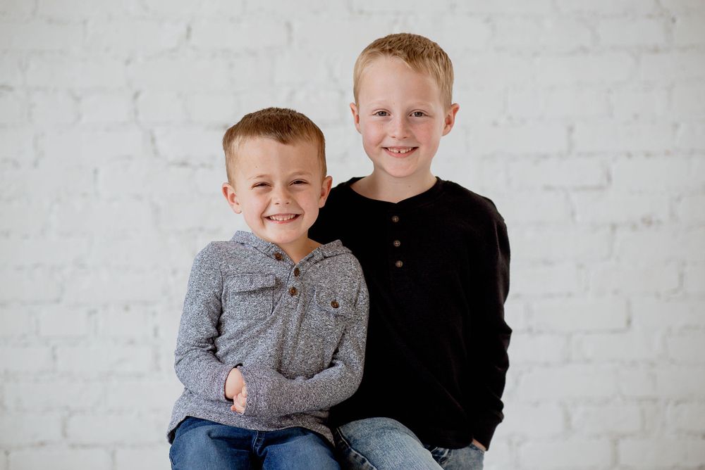 Two young children sit together smiling against a white brick wall, wearing casual sweaters.
