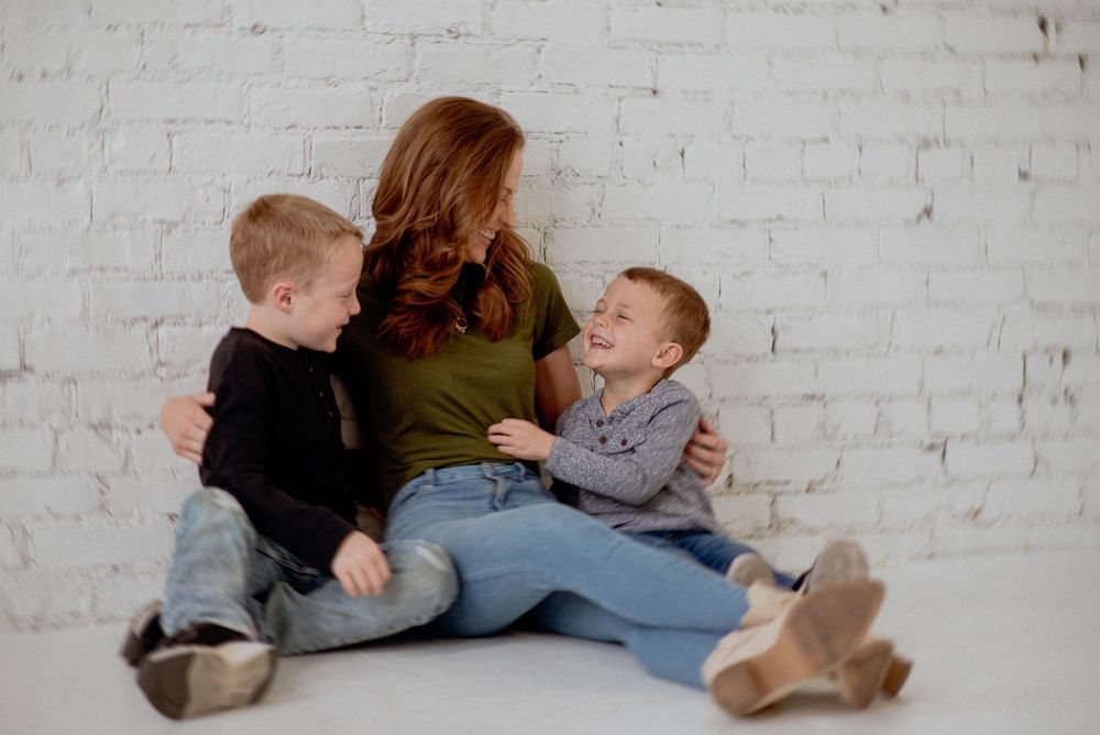 Three people sit together on floor sharing a joyful moment against a white brick wall backdrop.