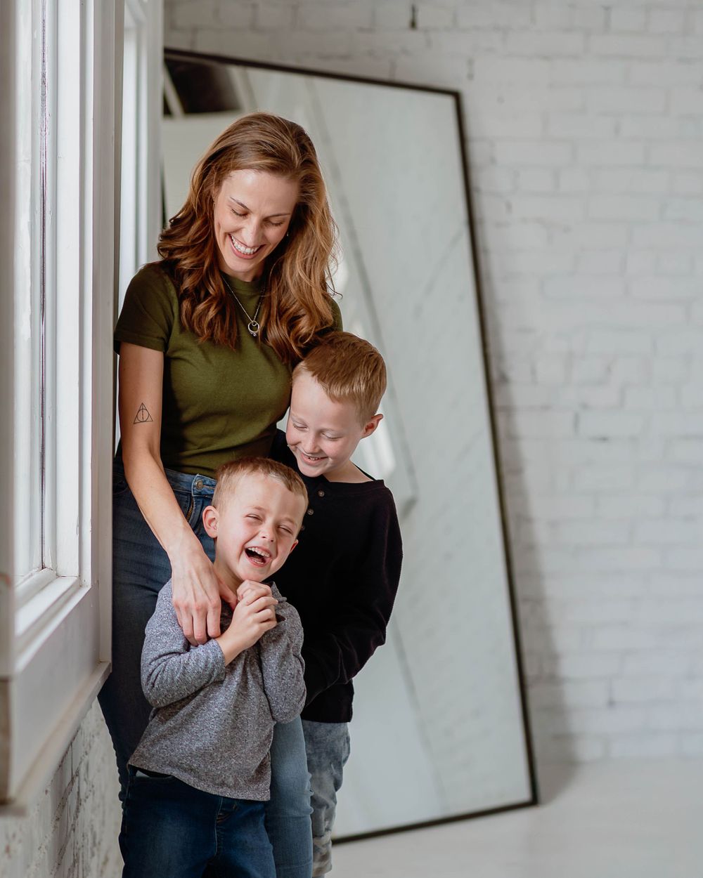 Family sharing tender moment and laughter by window with white brick wall and large mirror in background.
